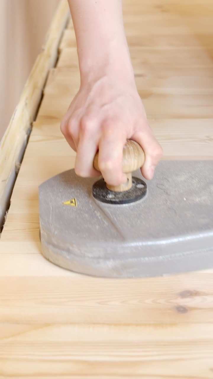 Hand guiding sander along narrow wood floor edge near white baseboard area, vertical closeup