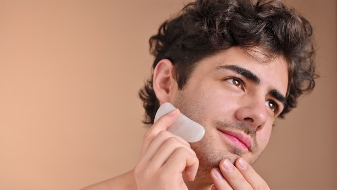 A young caucasian man with stubble beard is doing a facial massage using a Gua Sha. Slow motion