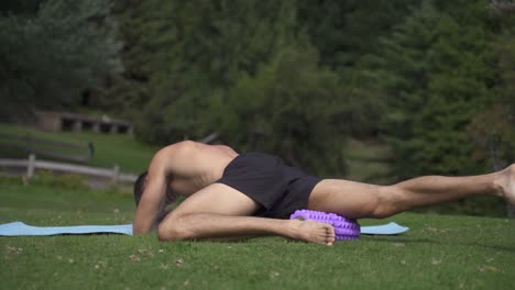 Young-caucasian-fit-attractive-male-exersicing-outdoors-Foam-roller-upper-legs-with-green-vegetation-background-slow-motion-60-fps