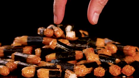 A hand selects a seaweed-wrapped roasted rice cracker from a pile under studio lighting, with a black background and smooth camera movement