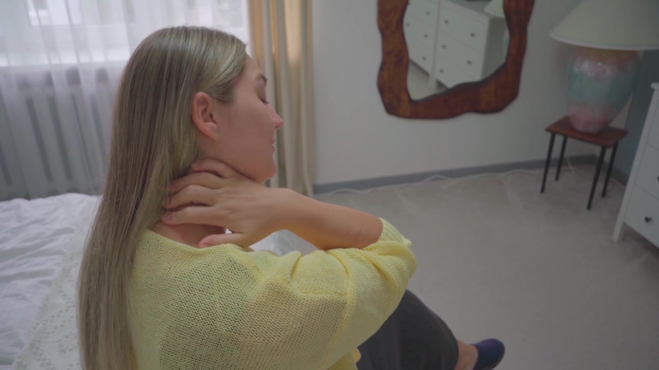 Ragazza bionda seduta sul bordo di un letto in una stanza semplice, che si tiene il collo con entrambe le mani in evidente disagio.