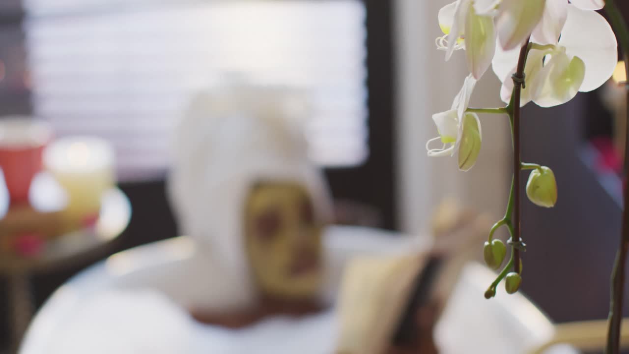 수건과 마스크를 착용 한 아프리카계 미국인 여성이 욕실에서 목욕하고 책을 읽고 있습니다.