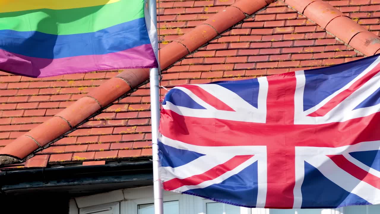 Close-up of rainbow and Union Jack flags waving in the wind with a red tiled rooftop background.
