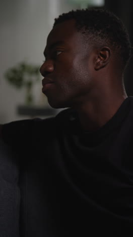 vertical video close up of unhappy and depressed young man sitting on sofa at home looking anxious and worried resting head on hand