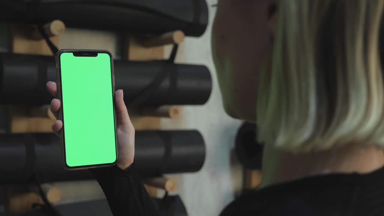 una joven atlética sostiene un teléfono inteligente en la mano, muestra una pantalla chromakey verde, está en el gimnasio