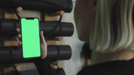 a young athletic woman holds a smartphone in her hand, shows a green chromakey screen, is in the gym