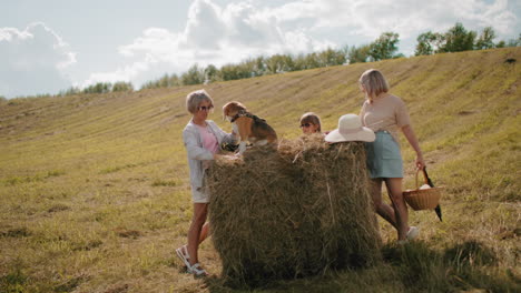 woman holding dog paw atop haystack while another woman carries picnic basket, a straw hat rests on hay, child peeks from behind, sunlit countryside with rolling hills and trees in background