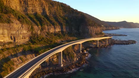 Camino-Curvilíneo-Del-Puente-Del-Acantilado-Del-Mar,-Una-Vista-De-Montañas-Majestuosas-Y-Aguas-Azules-Claras-Del-Mar-De-Tasman-En-Nsw,-Australia---Toma-Aérea