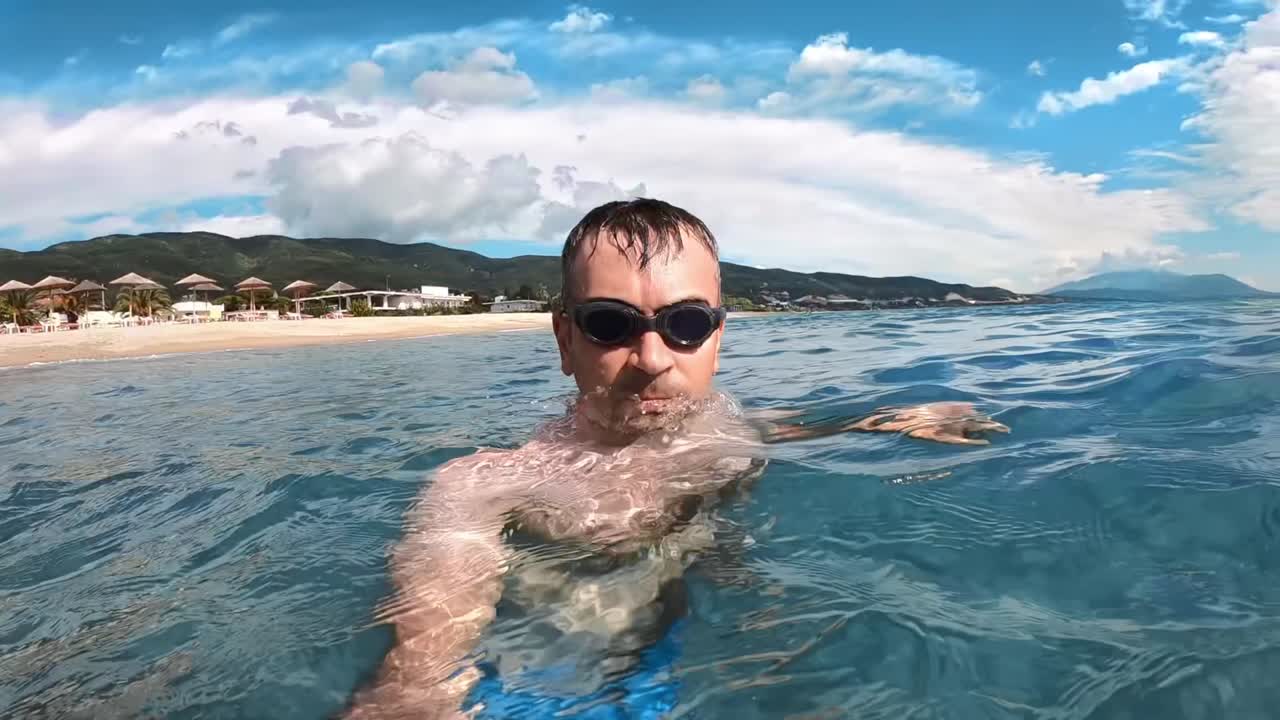 Man in swimming goggles in the water of the Aegean sea holding camera, nature on the background. Slow motion. Greece