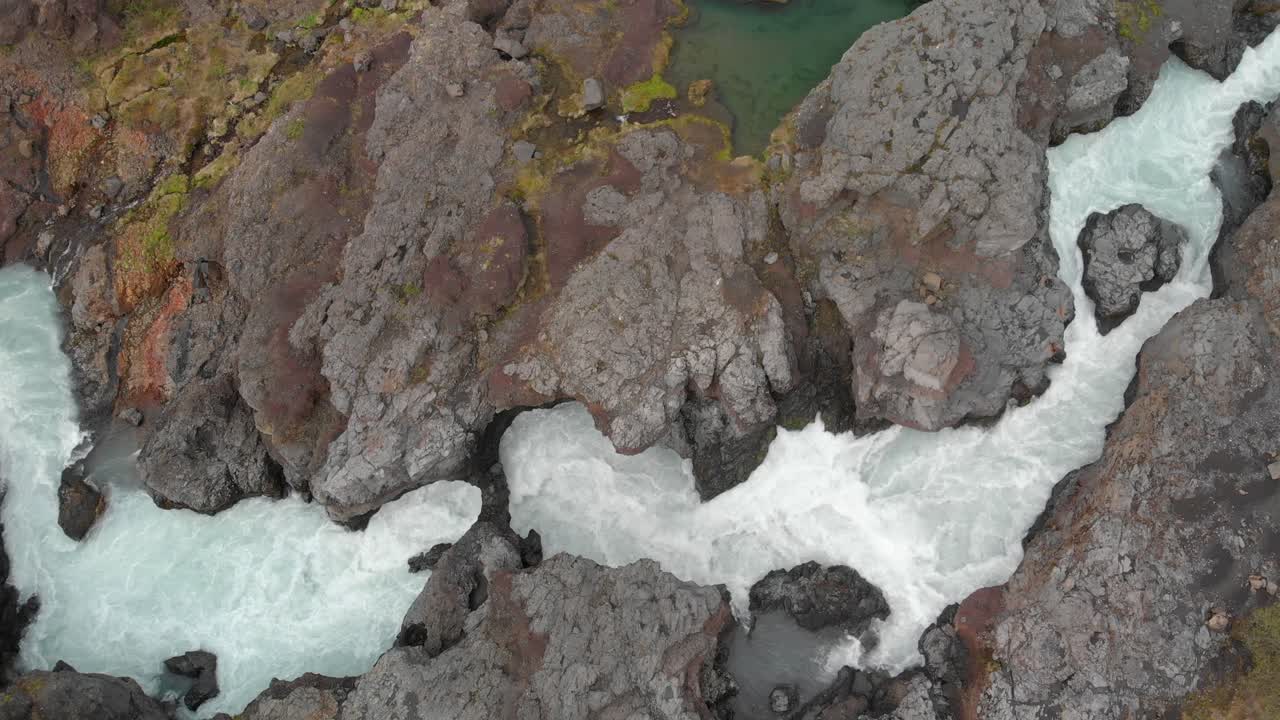 Flying with a drone up over the flowing river to waterfall Barnafossar surrounded by amazing rock formations and green landscape