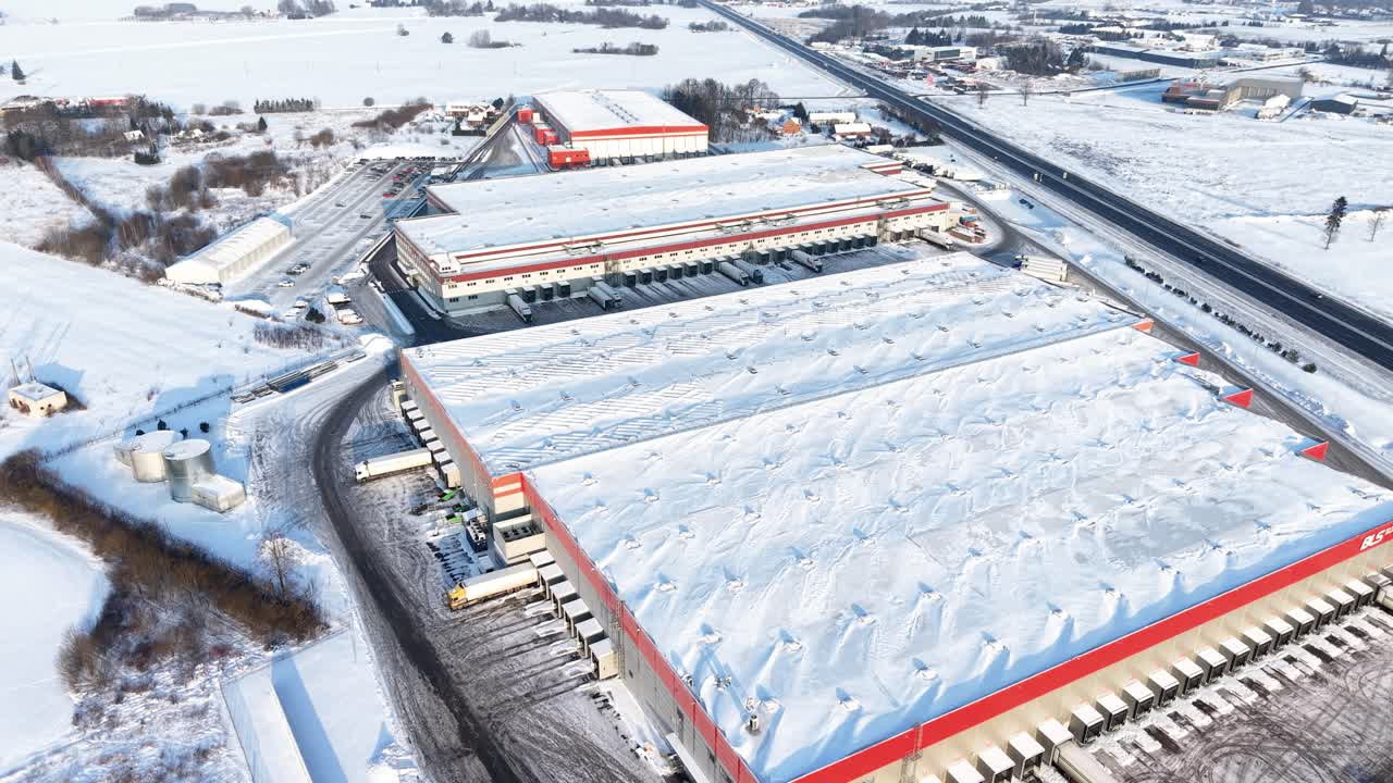 Drone video of a large industrial warehouse complex blanketed in snow, showcasing wide roofs and surrounded by truck parking. Captures the serene, cold atmosphere with clear blue sky