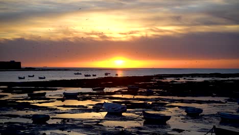 Stunning-beach-at-low-tide-with-fishing-boats-during-vibrant-orange-sunset