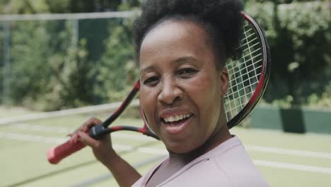 Portrait-of-happy-senior-african-american-woman-with-tennis-racket-at-tennis-court-in-slow-motion