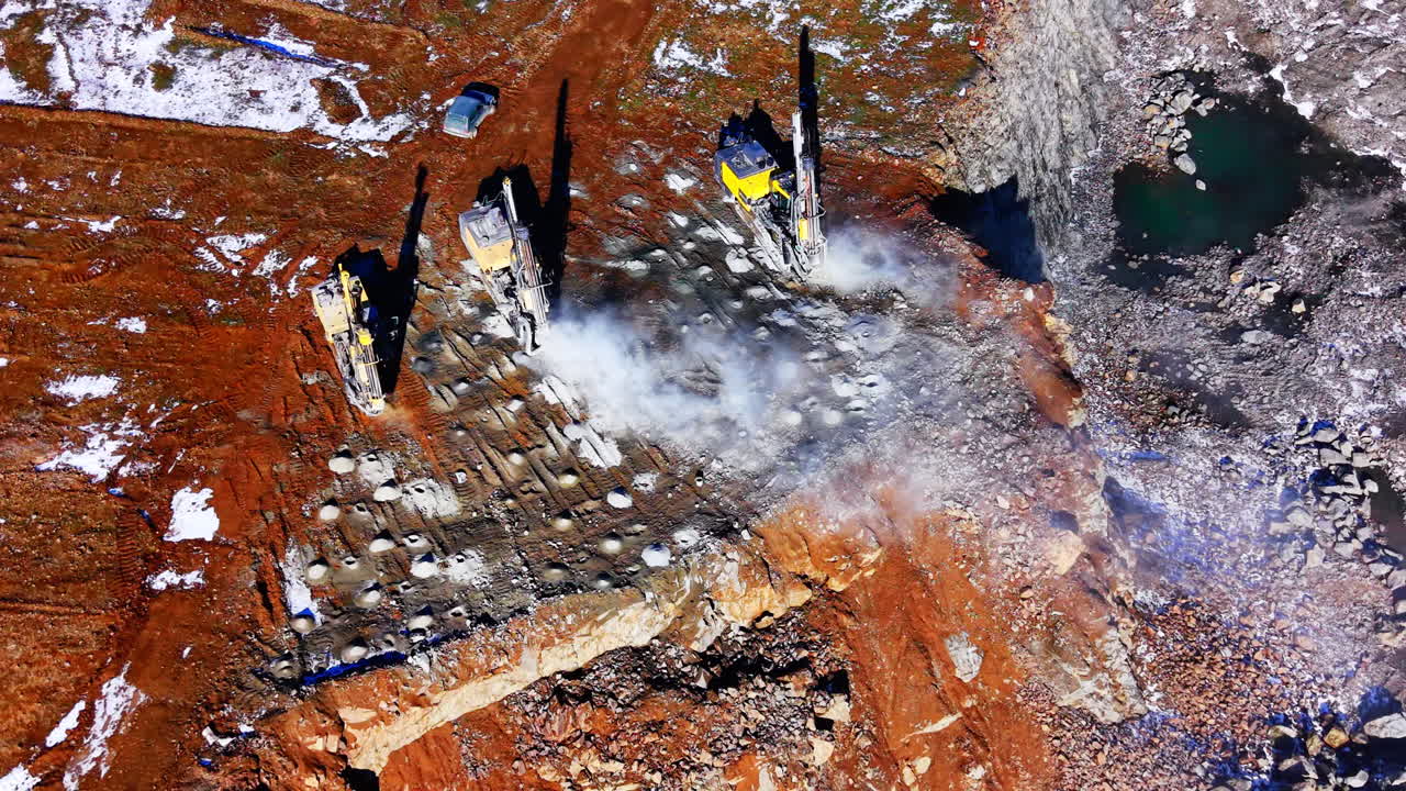Top-down luftfoto af tre kraftige boremakiner, der skaber huller i en stenet overflade på et mineområde med støv. Borerigge arbejder på klippeoverfladen i en åben mine.
