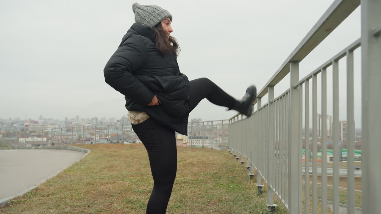 Şehir manzarası eşliğinde bir dans ısınması: Korkuluğa yüksek tekme atıp ardından oturarak esneme hareketleri yapan, şişme ceket ve örgü bere giymiş tek bir dansçı, bacak uzatma pratiği yapıyor. Kapalı şehir silüeti ve soluk kış ışığı altında neşeli bir sahne.