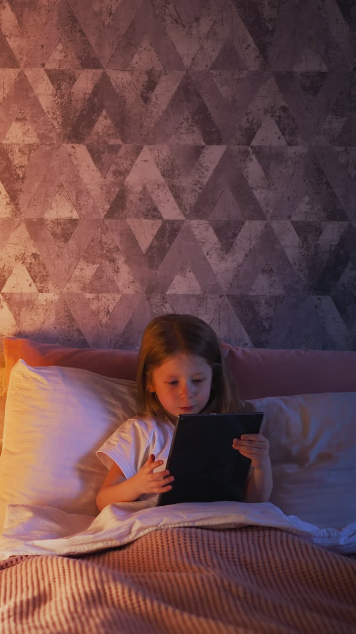 Addicted little kid plays video game on tablet resting in comfortable bed with soft pillows and plaid near toy house with glowing lamp slow motion