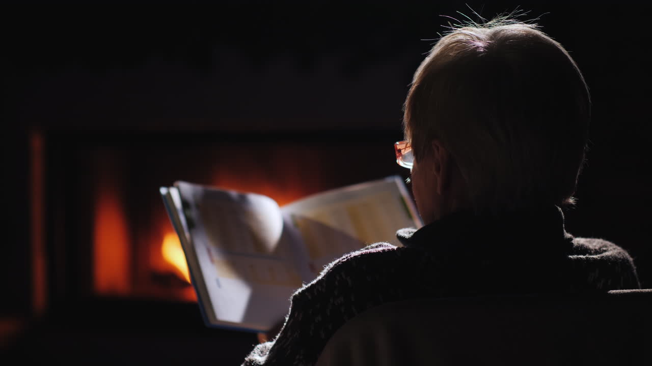 una anciana con gafas lee un libro junto a la chimenea