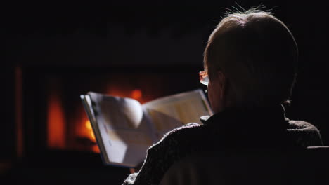 una anciana con gafas lee un libro junto a la chimenea