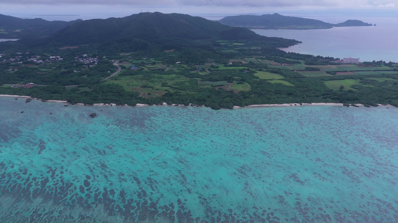 Inquadratura orizzontale che si allontana da un'angolazione obliqua elevata sopra acque costiere turchesi, barriere coralline e il litorale boscoso di Okinawa, Giappone