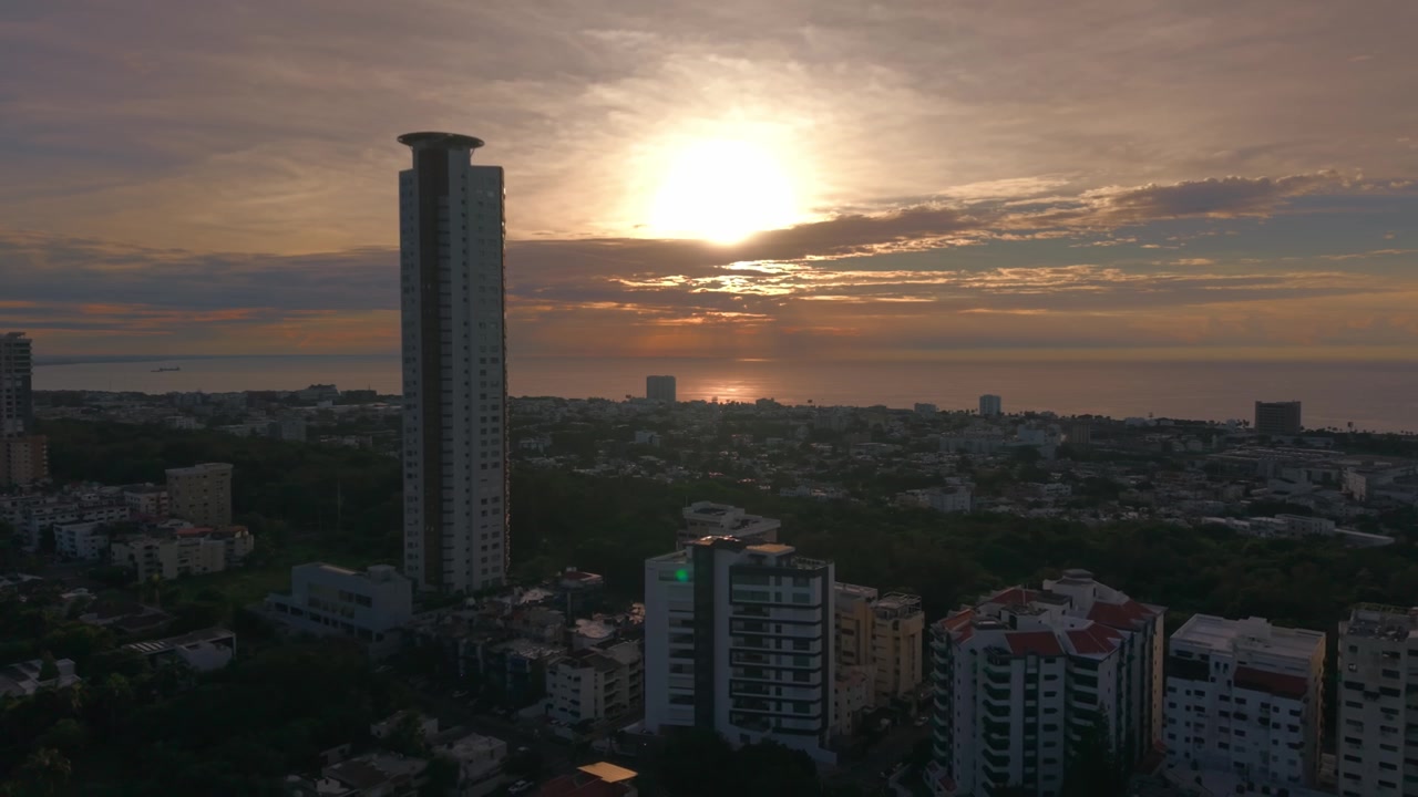 Imagem aérea de drone em ângulo alto, recuando sobre a paisagem urbana de Santo Domingo, exibindo o icônico arranha-céu Caney contra um nascer do sol dourado sobre o mar do Caribe