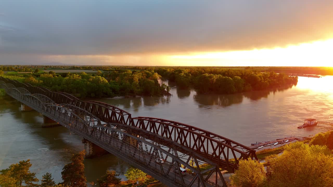 Ripresa aerea discendente con drone su uno storico ponte di ferro a Cremona che attraversa il fiume Po, con la luce dell'ora d'oro che illumina l'acqua, le rive lussureggianti e verdi e l'orizzonte lontano in Lombardia, Italia.