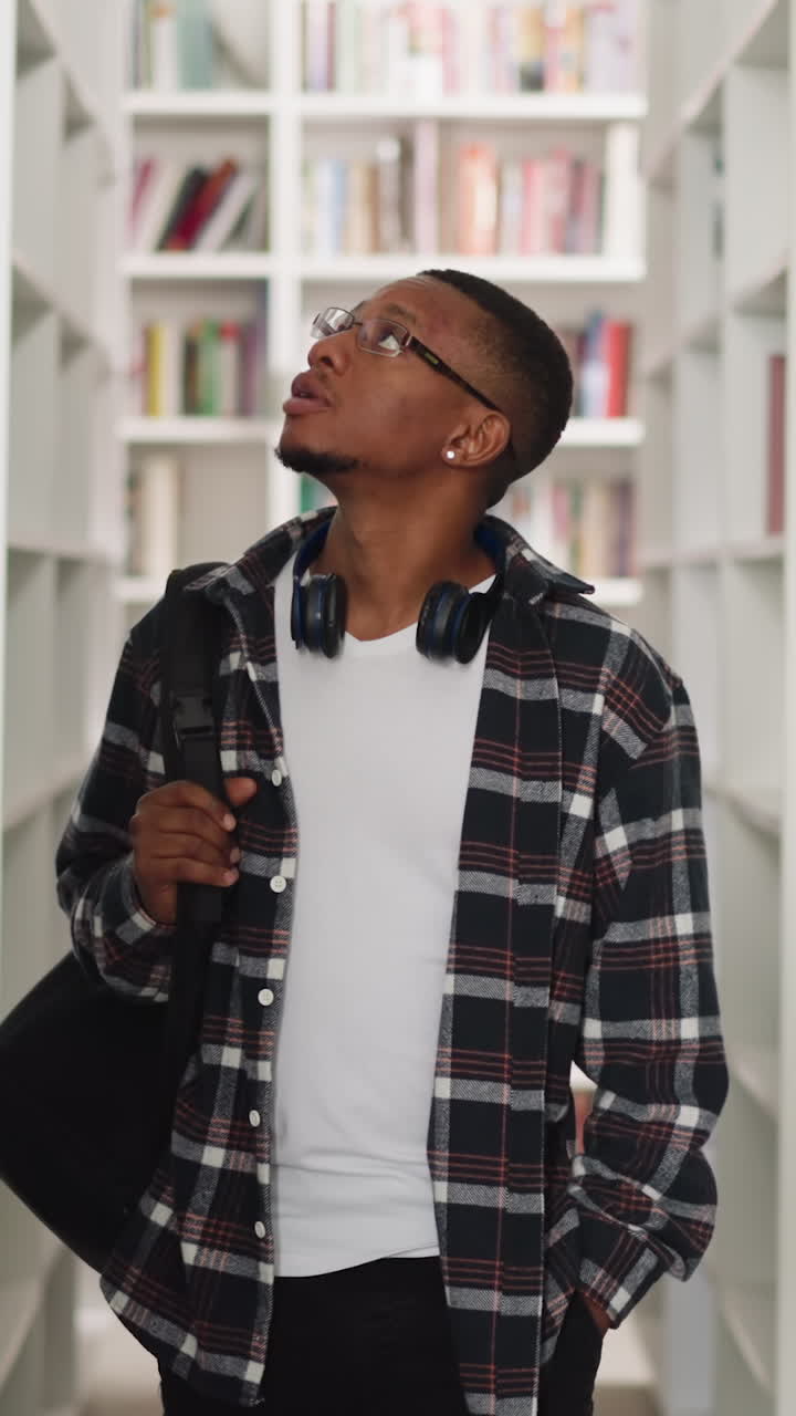 Young student looks around in college library. African American man in checkered shirt seeks textbooks in store. Informational sources storage