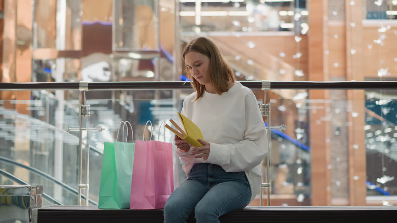 mujer joven sentada en un banco dentro de un centro comercial moderno leyendo un libro tomado de una bolsa de compras rosada, rodeada de un ambiente comercial colorido, barandillas de vidrio y escaleras mecánicas borrosas que se mueven hacia arriba