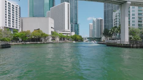 forwards tracking of motorboat group passing under bridge in modern city. miami river and downtown buildings on waterfronts. miami, usa