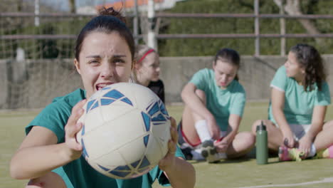 happy teenaged girl holding football in front of camera, while her teammates relax behind