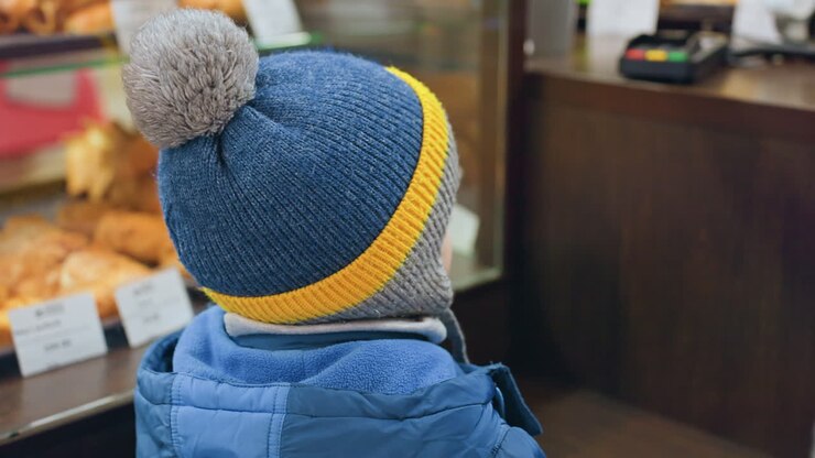 Mamma e figlio al bancone della pasticceria che scelgono un dolcetto, gesto affettuoso mentre la mano dell'adulto guida il bambino con il berretto blu e pompon, vetrina piena di croissant e panini, cartellini dei prezzi ed etichette visibili, atmosfera accogliente