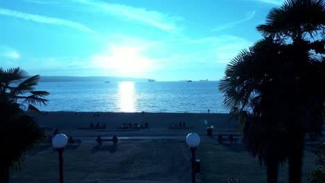 English-Bay-Vancouver-Bc-Frühe-Dämmerungsdrohne-Fliegt-In-Den-Eingang-Des-Strandbereichs,-Während-Sich-Menschen-Versammeln,-Um-Die-Sonnenuntergangsschatten-Von-Menschen-Von-Hinten-Zu-Beobachten,-Die-Sitzen-Und-Wandern-Und-Frachtboote-Im-Hafen-Beobachten