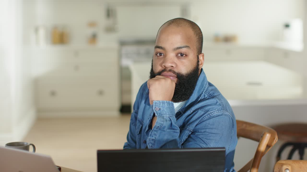 African American man sitting at kitchen table in denim shirt reading message on laptop smiling