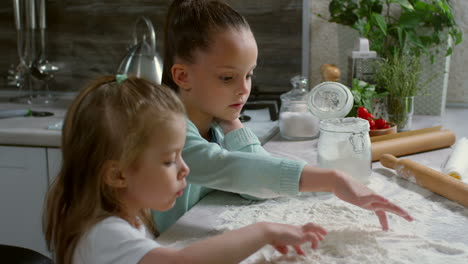 two little sisters of primary school age cooking in kitchen and drawing with fingers on floured surface