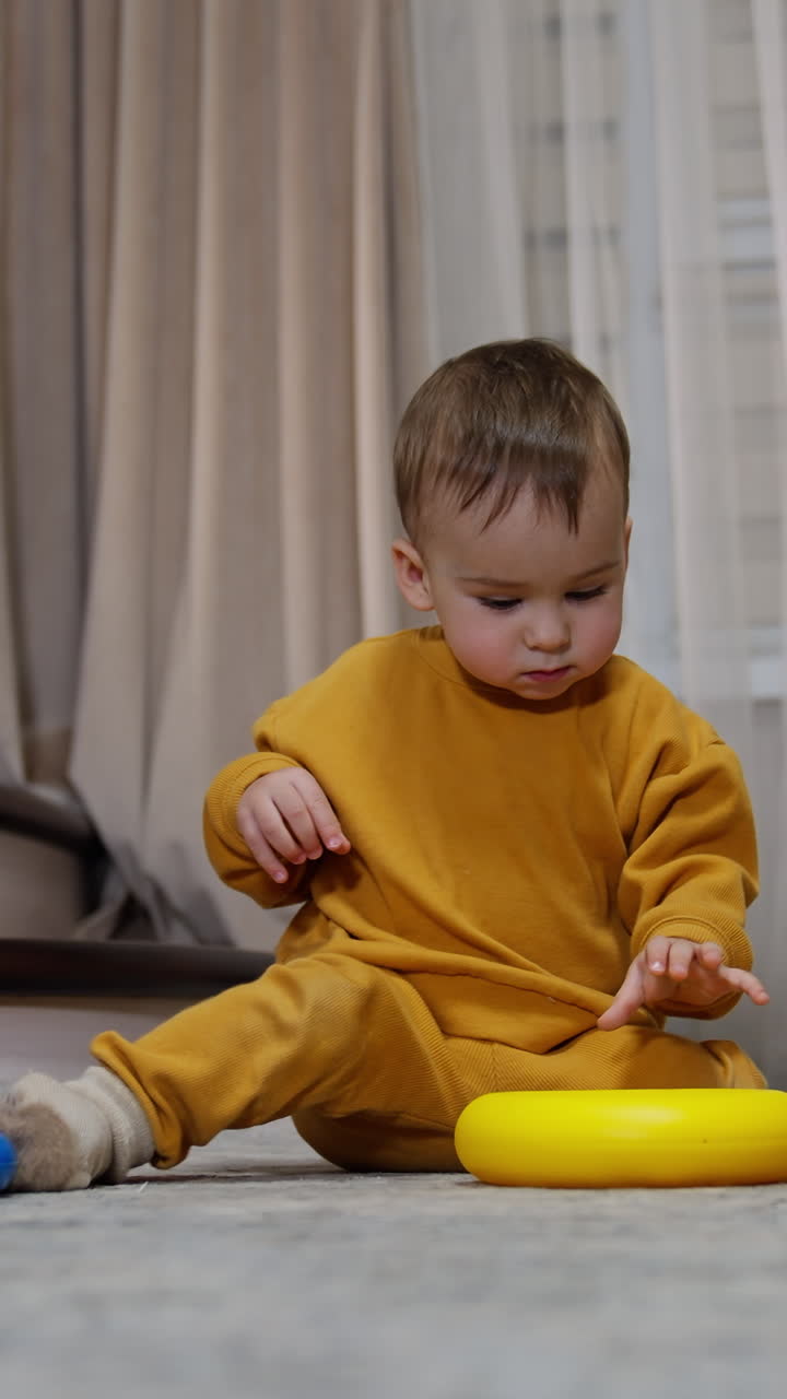 Baby boy in orange suit plays with a toy pyramid on the room floor. Little kid takes one piece and heads to the stairs. Vertical video
