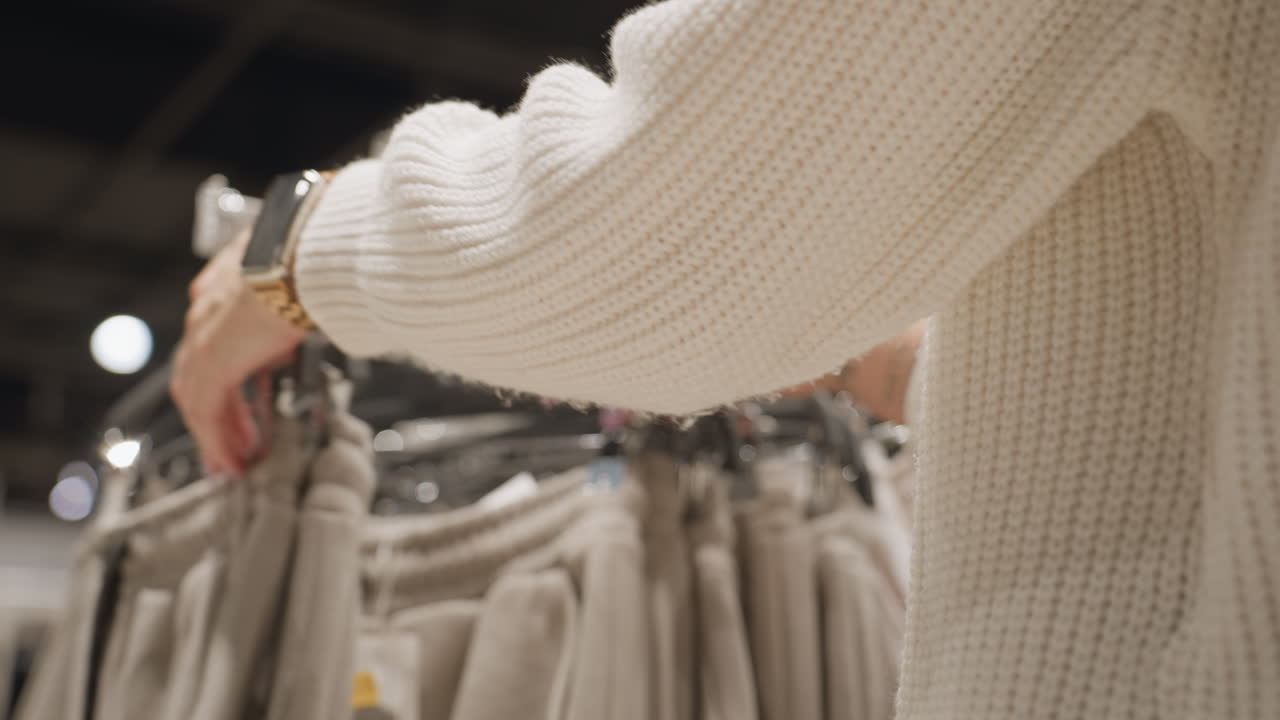 Housewife hands in chunky knit sweater glide over beige joggers on metal rack inside clothing store, brushing waistbands assessing texture, size, choosing comfy loungewear while aisle lights glow