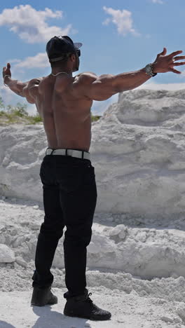 muscular dark skinned man without shirt stands on white hill. shirtless african athlete stands with stretched arms among summer nature. sportsman in the mountains. rear view. vertical video