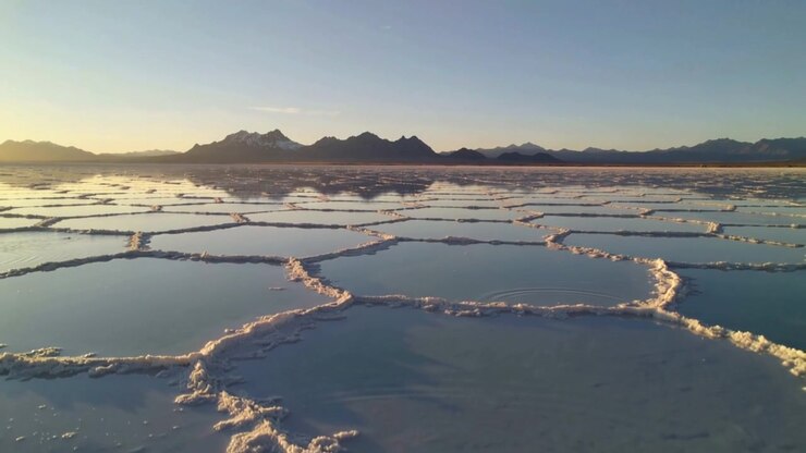 Una telecamera drone che scivola rivela una crosta di sale poligonale in una salina, catturando i riflessi delle montagne.