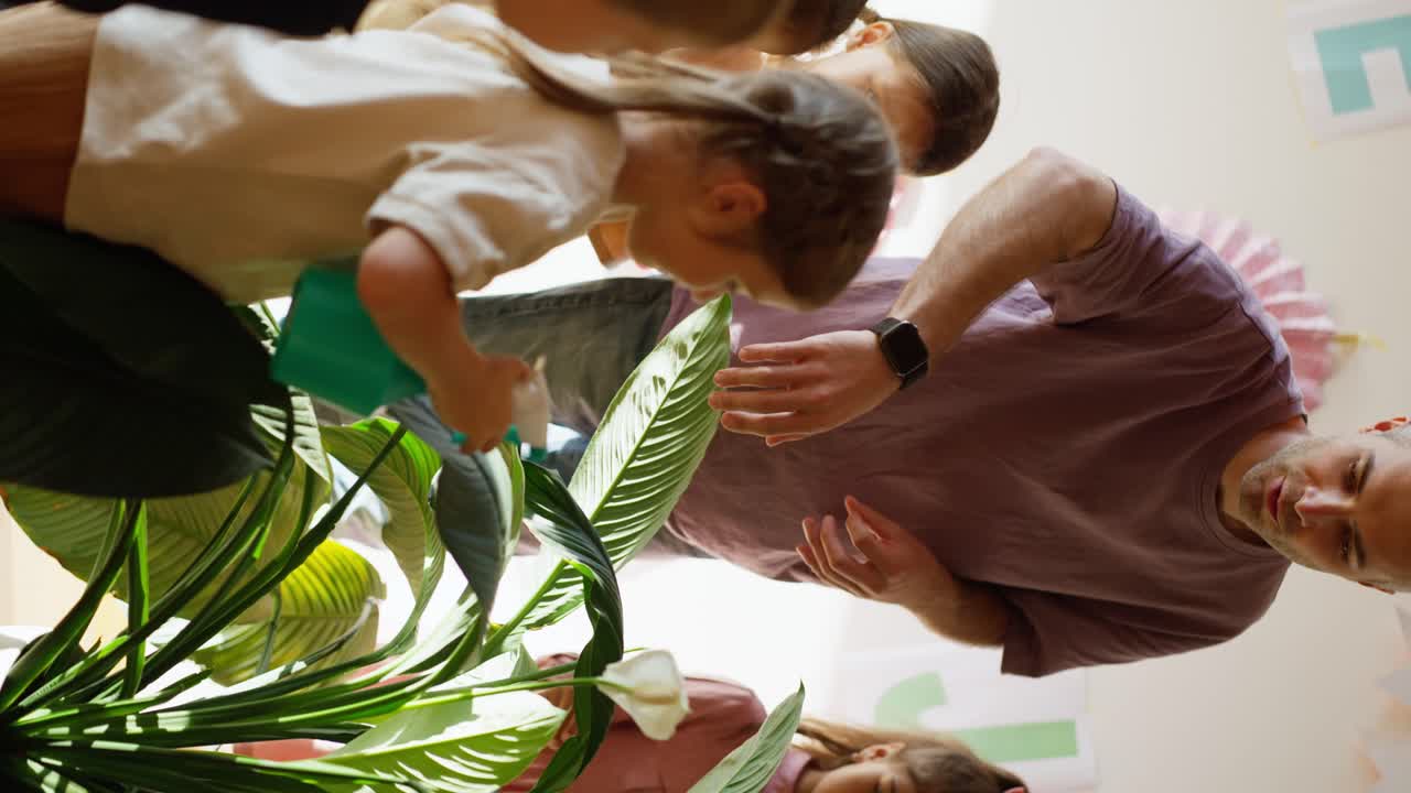 video vertical de un hombre con cabello gris en una camiseta púrpura enseñando a los niños en edad preescolar cómo regar correctamente las flores usando una botella de spray en una sala de club para preparar a los niños para la escuela