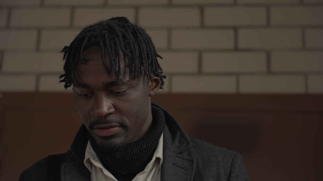 close de um homem de cabelo natural com casaco cor de carvão olhando para baixo enquanto desce as escadas, plano de fundo de tijolos levemente borrado, alça de ombro visível, camadas de inverno, movimento implícito durante o passo