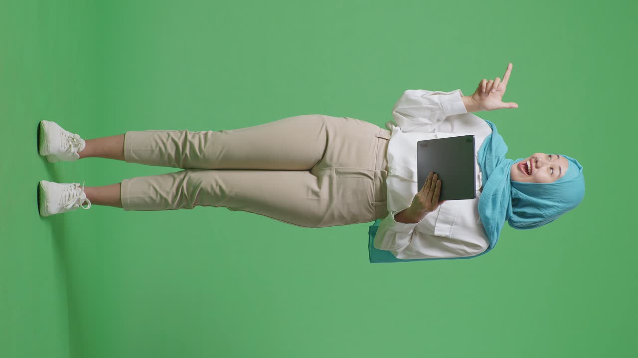 cuerpo lleno de mujer musulmana asiática sonriendo y usando la tableta luego apuntando a un lado mientras está de pie en la pantalla verde estudio de fondo