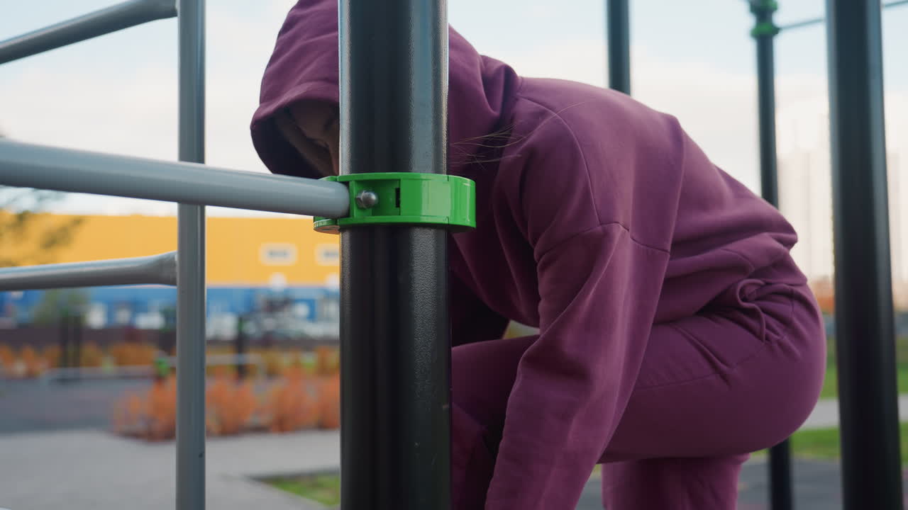 Wooden bench surface at outdoor fitness area with blurred view of active woman in hoodie tying sneaker lace beside calisthenics bars with gym bag resting on grass under urban skyline