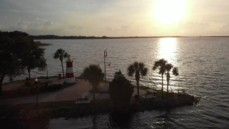 Bird-crossing-sunset-Aerial-drone-wide-shot-of-marina-boat-dock-with-lighthouse-at-the-bay-palm-trees