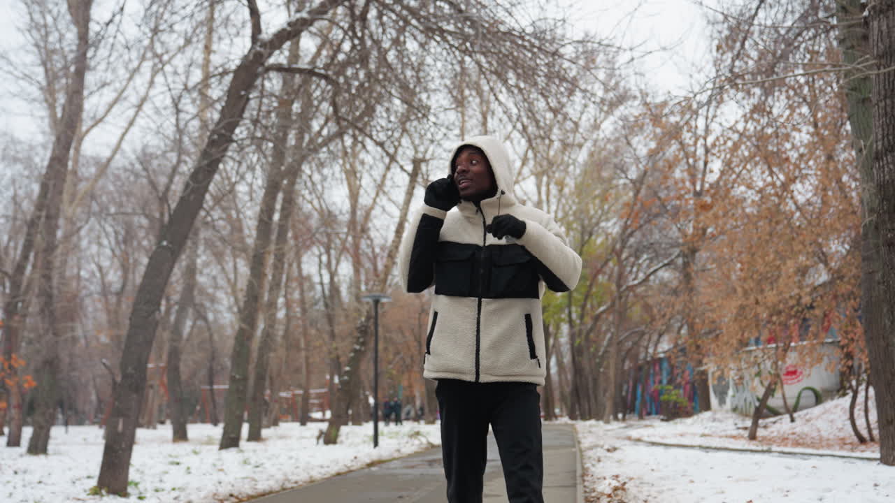 Homme africain parlant au téléphone avec quelqu'un en se promenant dans un parc en hiver, il porte un sweat à capuche blanc et des gants, avec des arbres, de la neige et des bâtiments flous en arrière-plan.