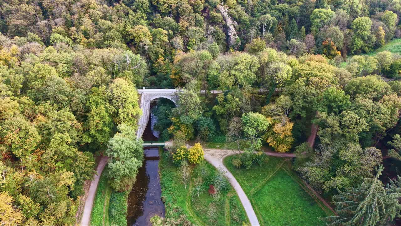 Old stone bridge over river in autumn forest, Forêt de Concise, Saint-Berthevin, France. Aerial drone forward