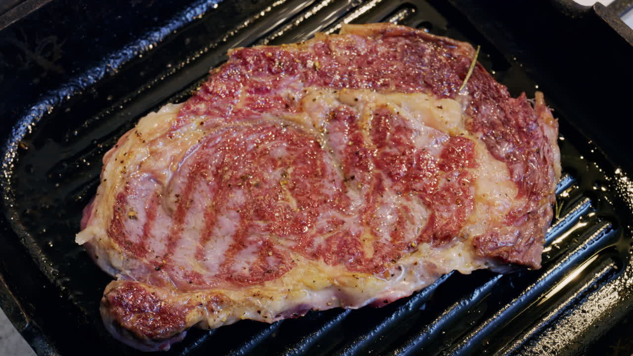 Mantequilla derritiéndose sobre un bistec sazonado cocinándose en una sartén grill caliente con hierbas de romero