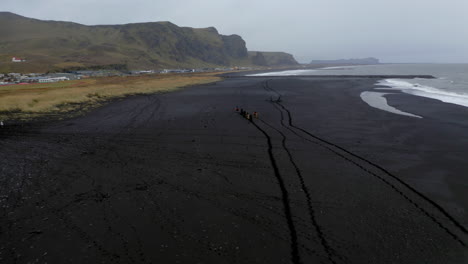 aerial: flying towards group of tourists riding horses on the black sand beach of vik in iceland