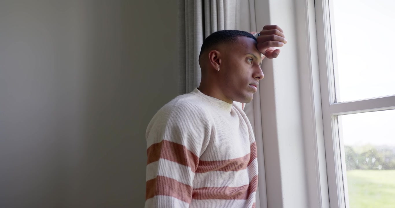 African American man standing by window in striped sweater raising right hand to brow reflecting
