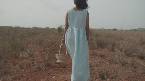 cinematic slow motion dolly shot of a young indian woman in light blue dress, black hair and a wood basket in her hands walking straight on a dry and dusty field