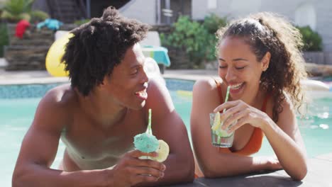 Two-happy-diverse-male-and-female-friends-toasting-with-drinks-in-swimming-pool