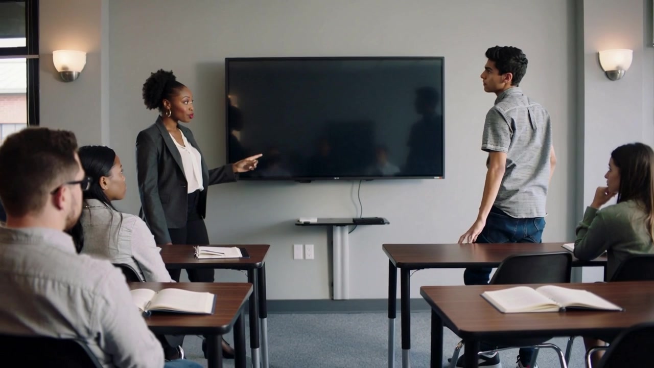 Pointing teacher in blazer calling grey-shirted student forward to monitor at classroom front